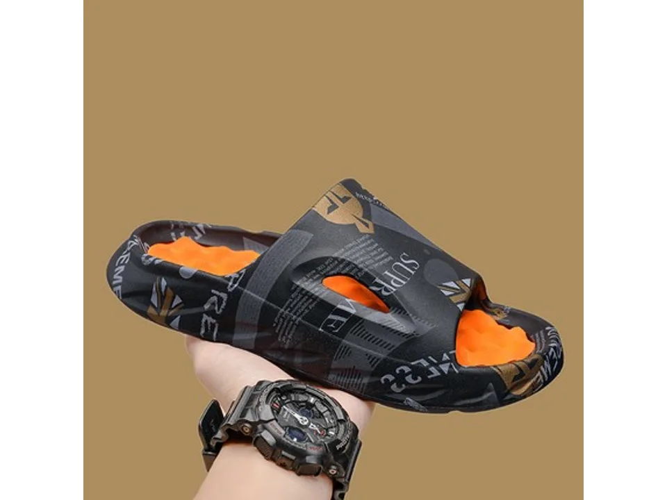 A black slide sandal with a graphic print and orange textured footbed, held in a hand.