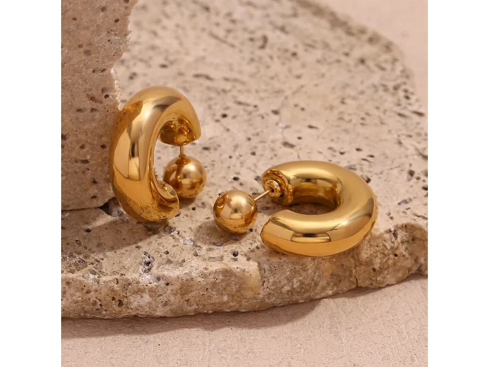 A pair of gold-tone hollow tube ball stud earrings resting on a textured stone surface.