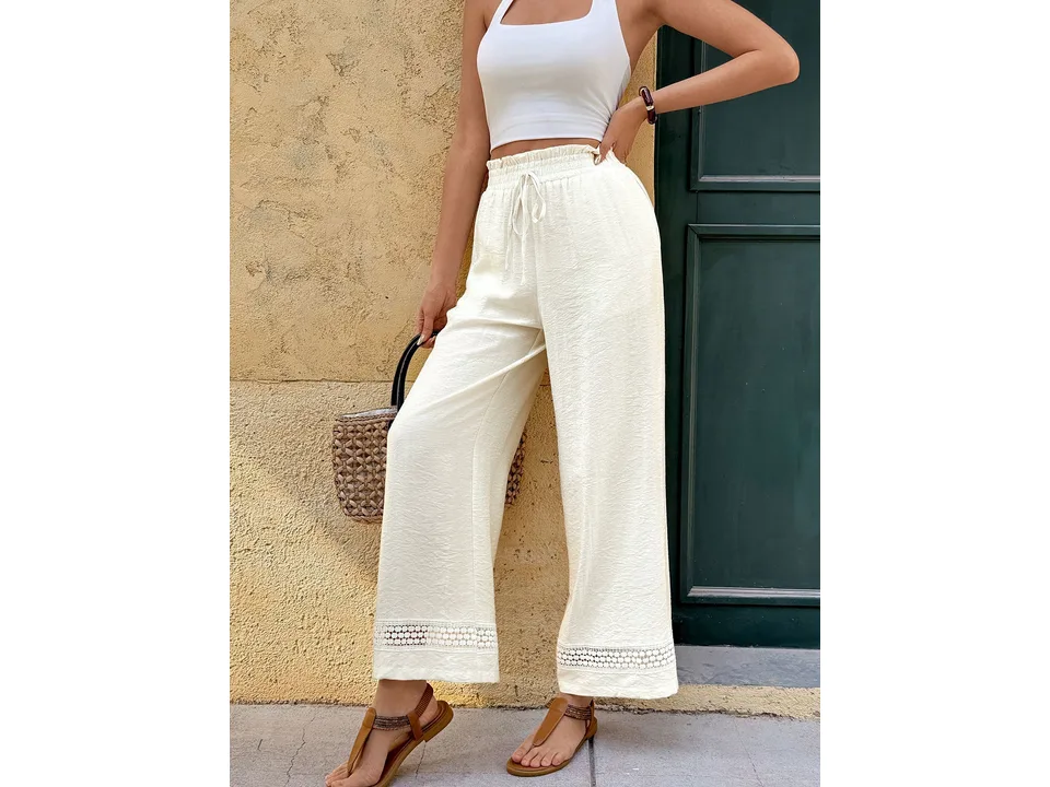 A woman wearing white wide-leg pants with lace trim, standing against a textured wall.