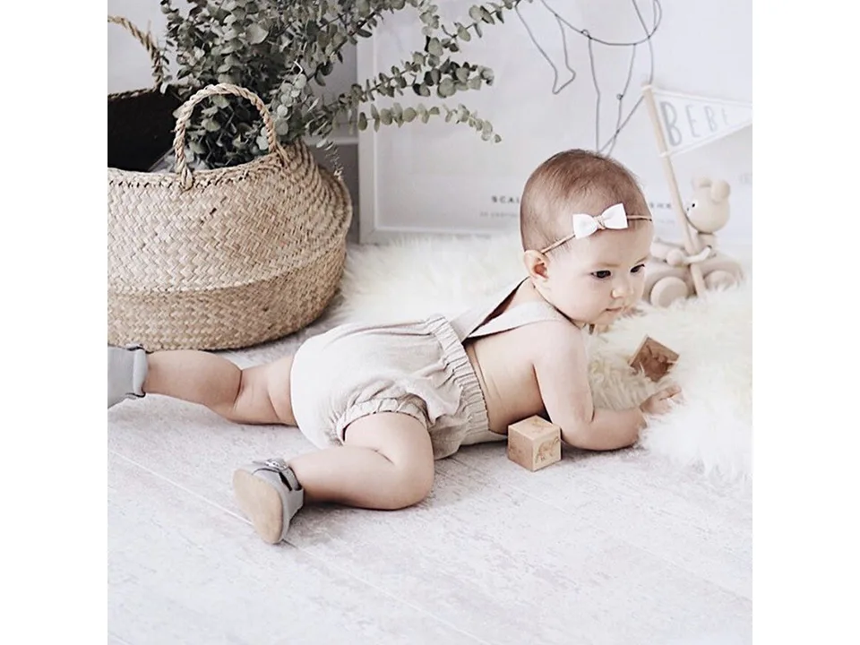 A baby lying on a rug wearing a beige linen suspender romper.