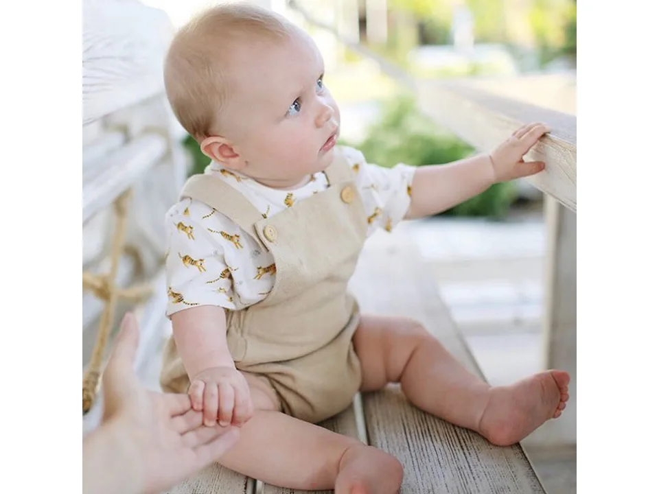 A baby sitting on a bench wearing a beige linen suspender romper with a tiger print shirt.