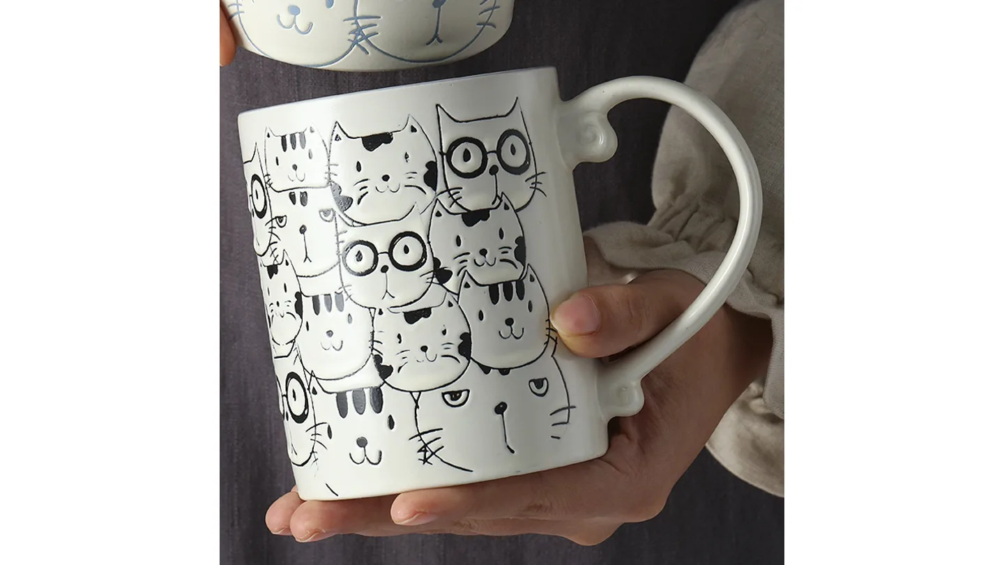 A person holding a white ceramic mug with a pattern of black and white cat faces.