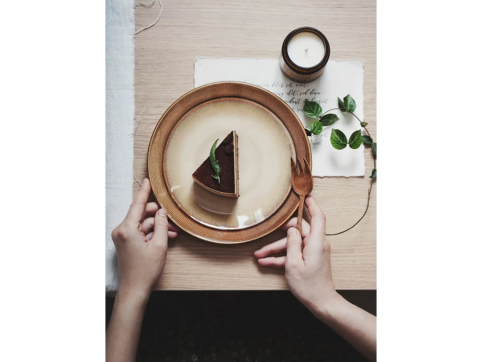 Hands holding a ceramic dish with a slice of cake on a wooden table.