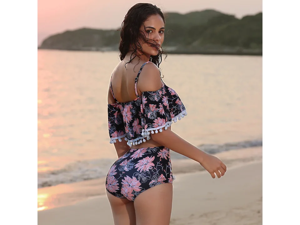 Model wearing a floral high-waist bikini with pom-pom trim, viewed from the back on a beach.