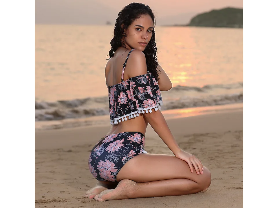 Model kneeling on the beach wearing a floral two-piece swimsuit with pom-pom details.