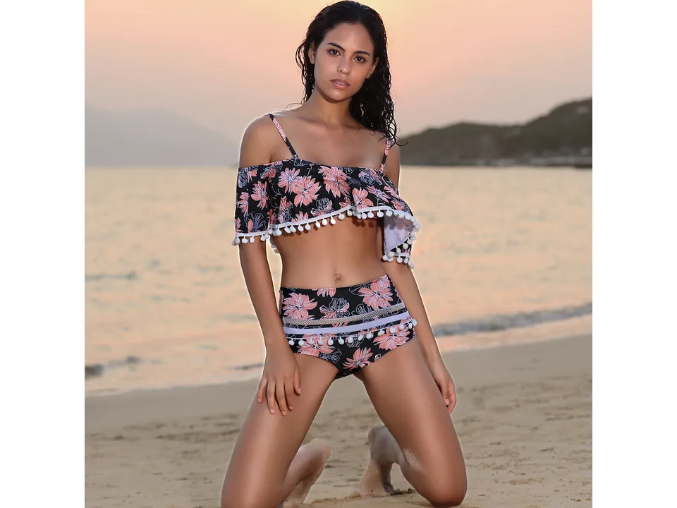 Model kneeling on the beach wearing a floral high-waist bikini with ruffled top and pom-pom trim.