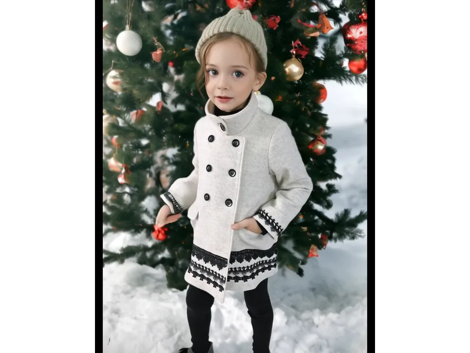 A young girl wearing a grey wool coat with black lace trim standing in front of a Christmas tree.