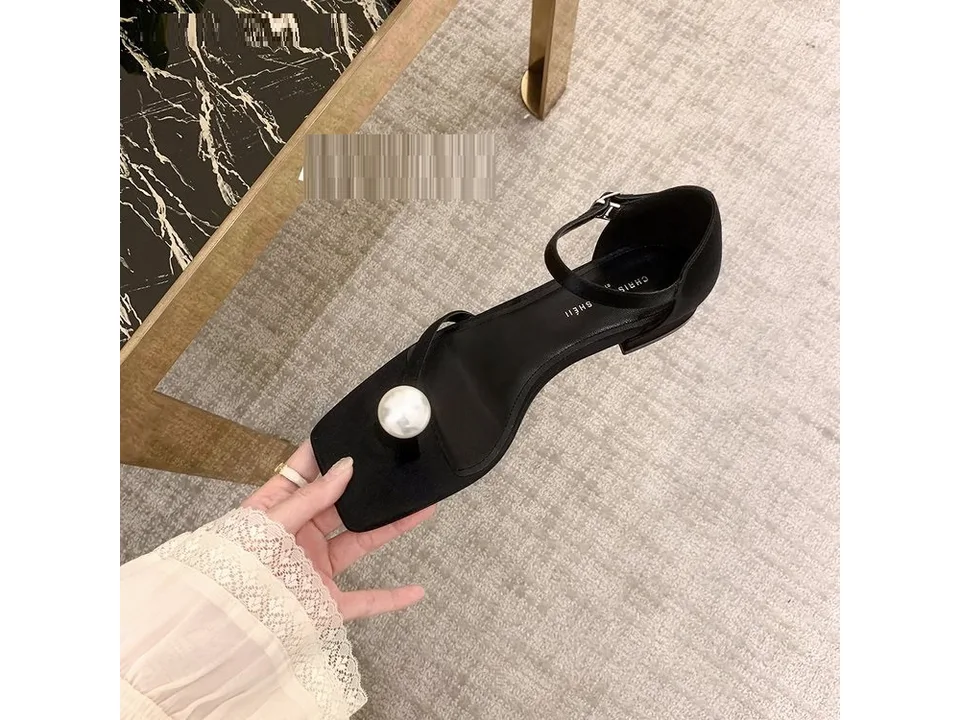 A black square-toe sandal with a large pearl detail held in a hand.