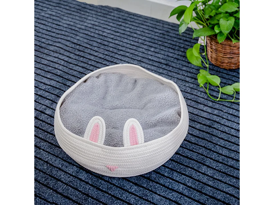 White round woven pet bed with pink bunny ears and a soft grey cushion, placed on a dark textured surface.