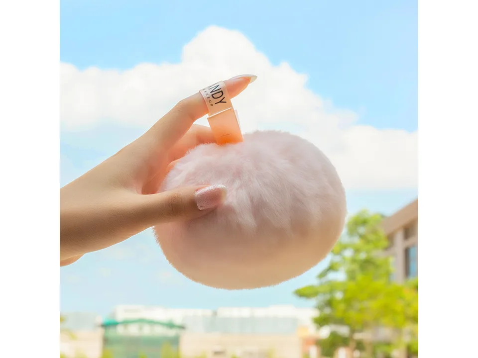 A hand holding the fluffy pink powder puff against a blue sky background.