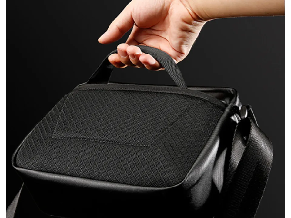 A hand holding the top handle of the black crossbody bag.