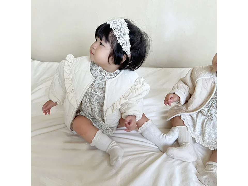 A baby sitting on a bed wearing the cream quilted jacket.