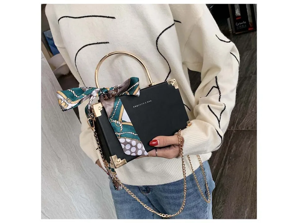 A person holding a black box-style handbag with a decorative scarf and gold chain strap.