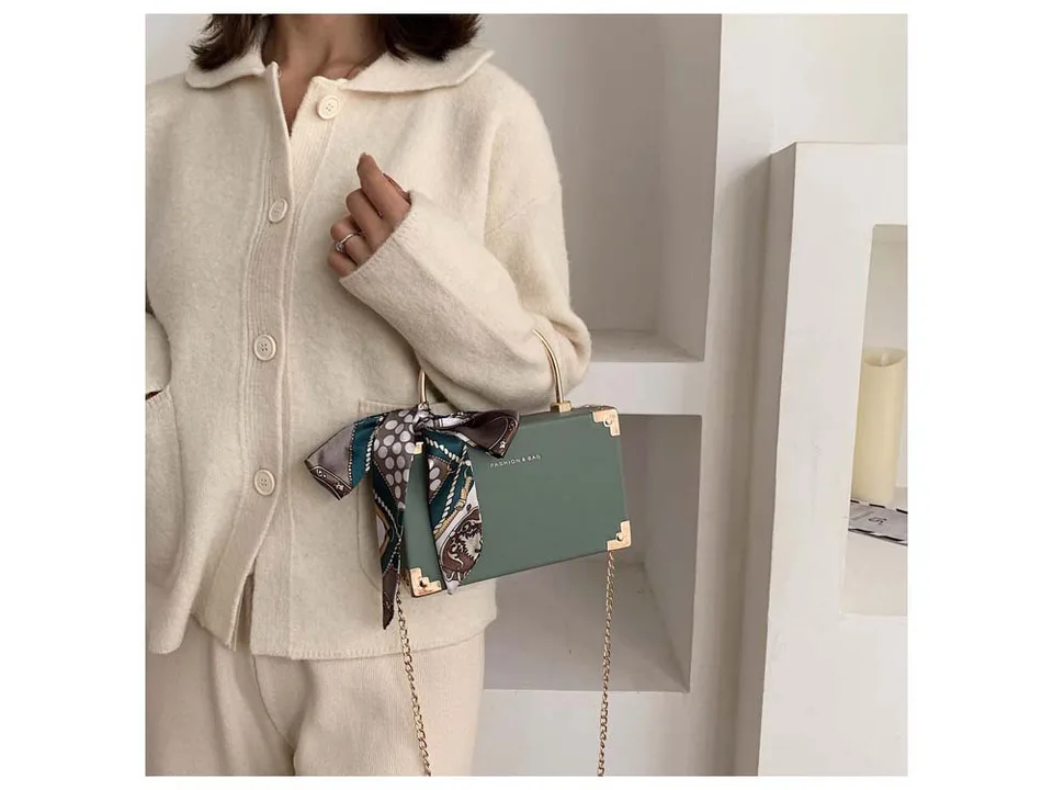A person holding a sage green box-style handbag with a decorative scarf and gold chain strap.