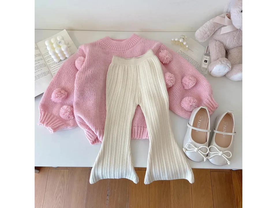 A pink pom-pom sweater, cream flared pants, and white shoes laid out on a table.