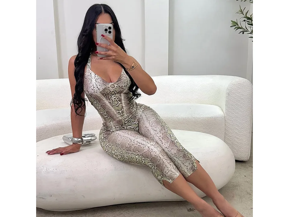 A woman wearing a snake-print halter neck jumpsuit while sitting on a white couch.