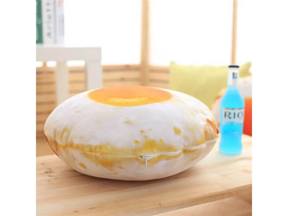A fried egg shaped plush pillow sitting on a wooden table next to a blue bottle.