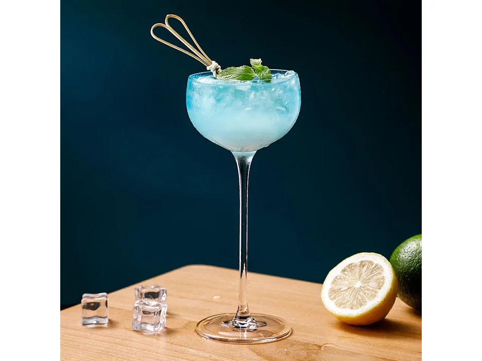 A stemmed cocktail glass filled with a blue drink, garnished with mint and a cocktail pick, on a wooden surface.