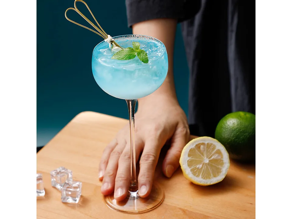 A hand holding a stemmed cocktail glass filled with a blue drink, garnished with mint and a cocktail pick.