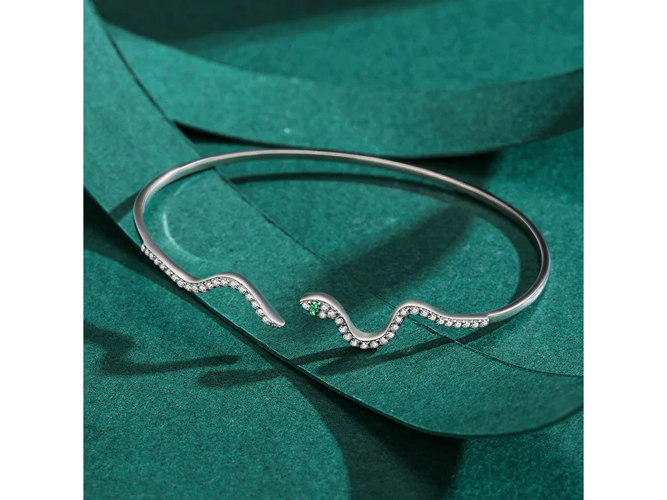 A close-up of a silver snake-shaped open cuff bracelet with clear stones and green eyes.