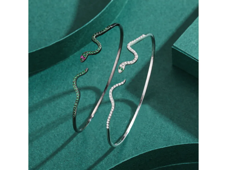 Two snake-shaped open cuff bracelets, one silver with clear stones and one dark metal with green stones.