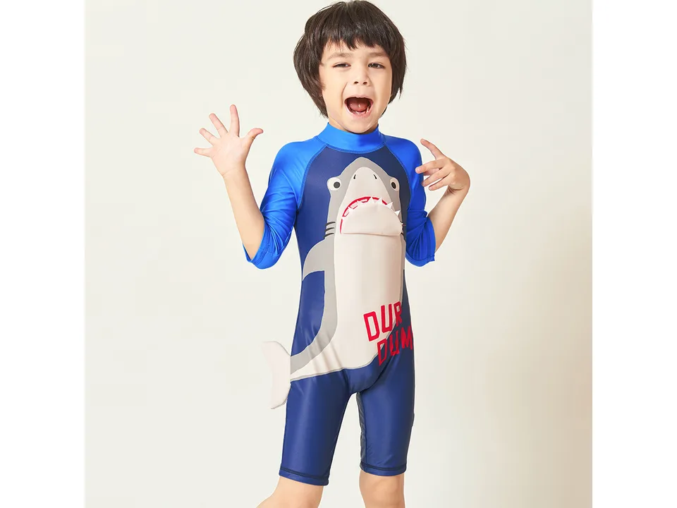 A young boy posing in a blue shark-themed one-piece swimsuit.
