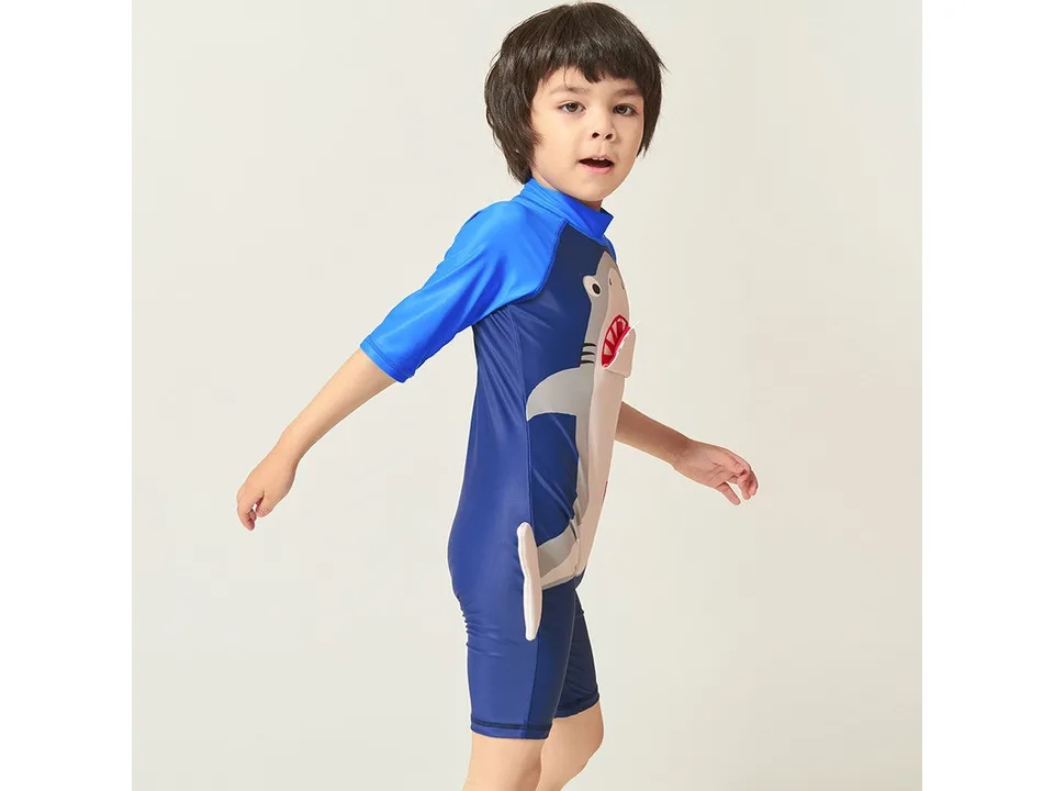 A side view of a young boy wearing a blue shark-themed one-piece swimsuit.