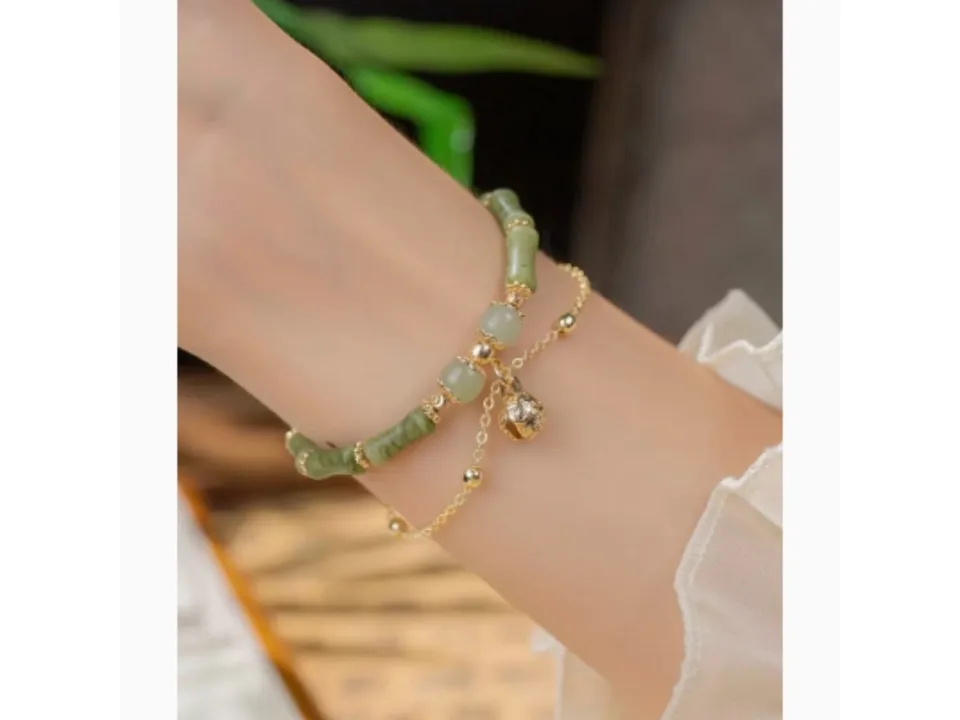 A green stone beaded bracelet with a gold chain and bell charm worn on a wrist.