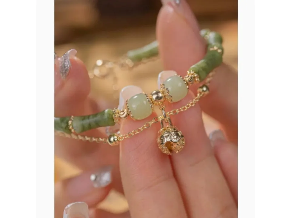 Close-up of a green stone beaded bracelet with a gold bell charm held by fingers.