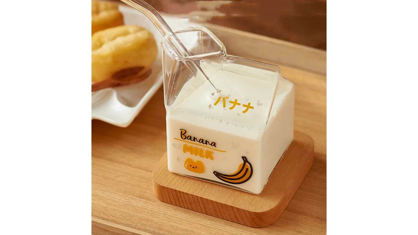 A square glass milk carton mug filled with milk, sitting on a wooden coaster.