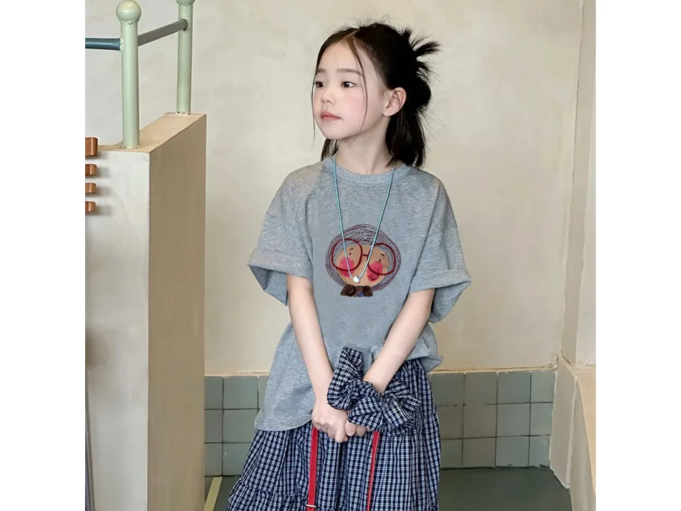 A young girl wearing a grey short-sleeved t-shirt with a cartoon face graphic.
