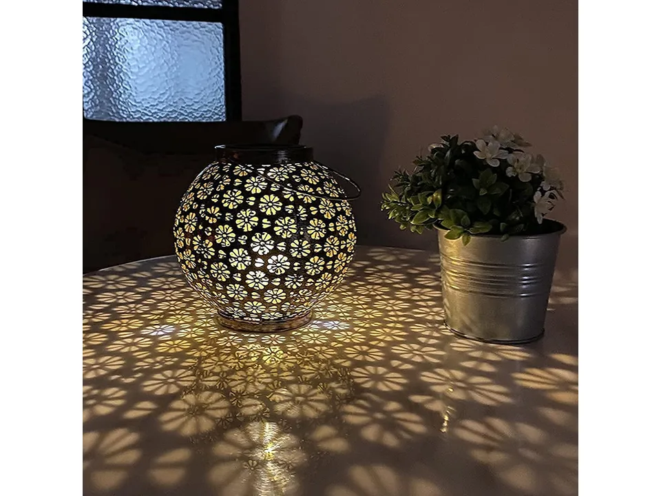 Solar lantern glowing on a table next to a small potted plant, casting flower-shaped shadows.