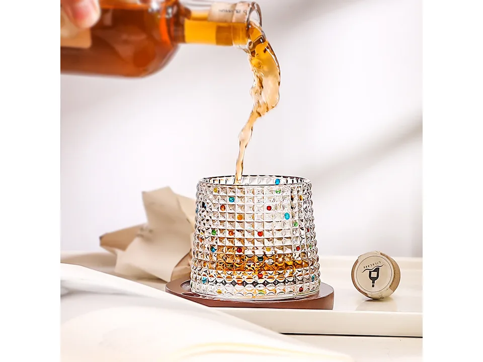 A textured glass tumbler on a wooden coaster being filled with a drink from a bottle.