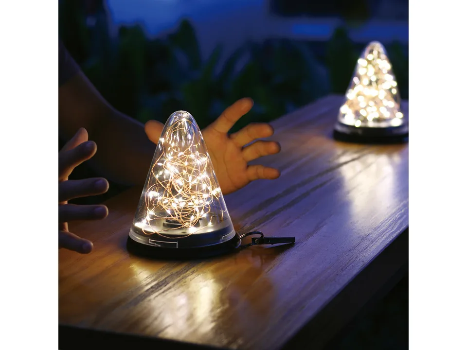 Solar cone light glowing on a wooden table with hands nearby for scale.