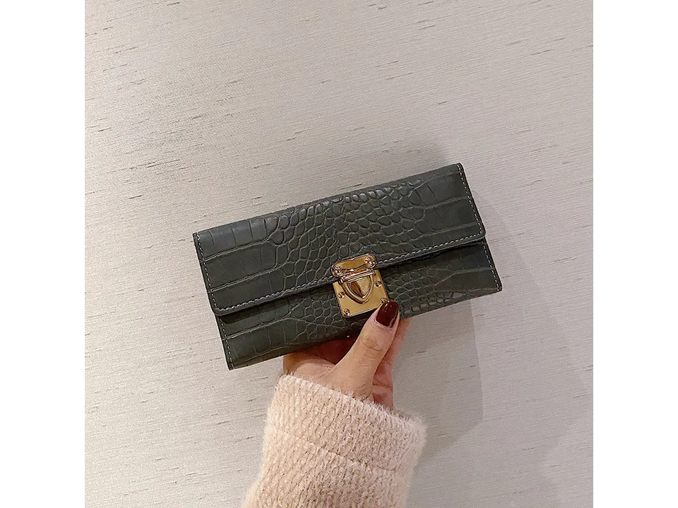 A grey crocodile pattern long wallet with a gold lock closure held in a hand.