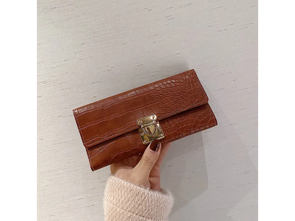 A brown crocodile pattern long wallet with a gold lock closure held in a hand.