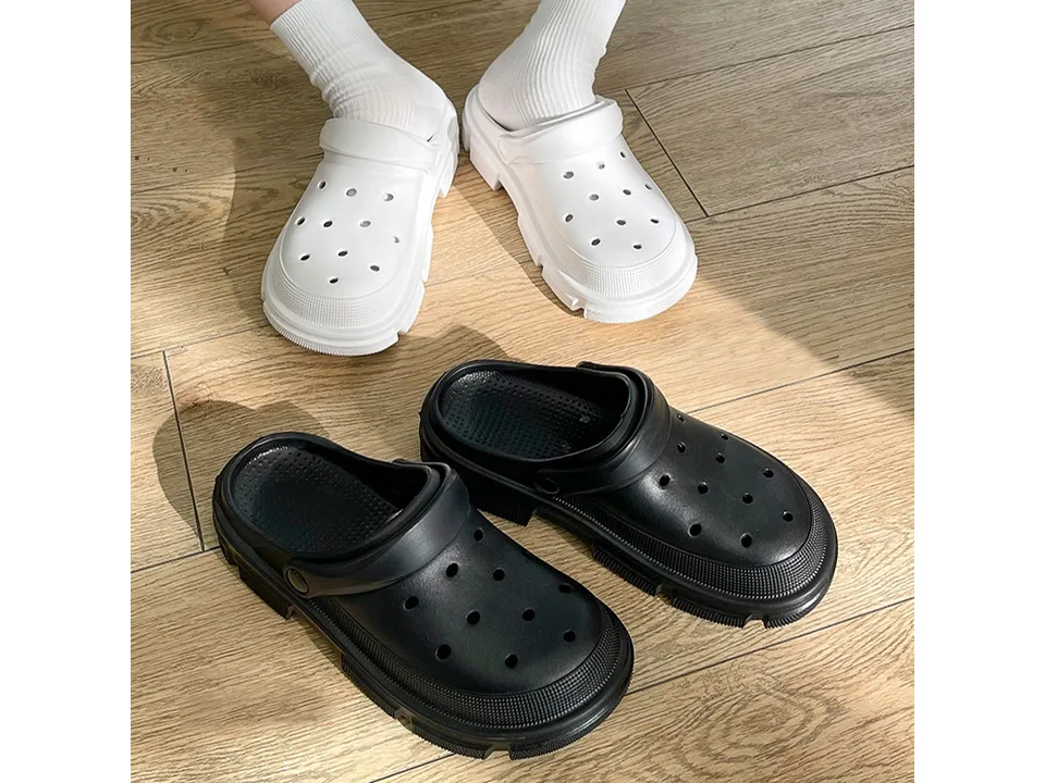 A person wearing white EVA clogs and a pair of black EVA clogs on a wooden floor.
