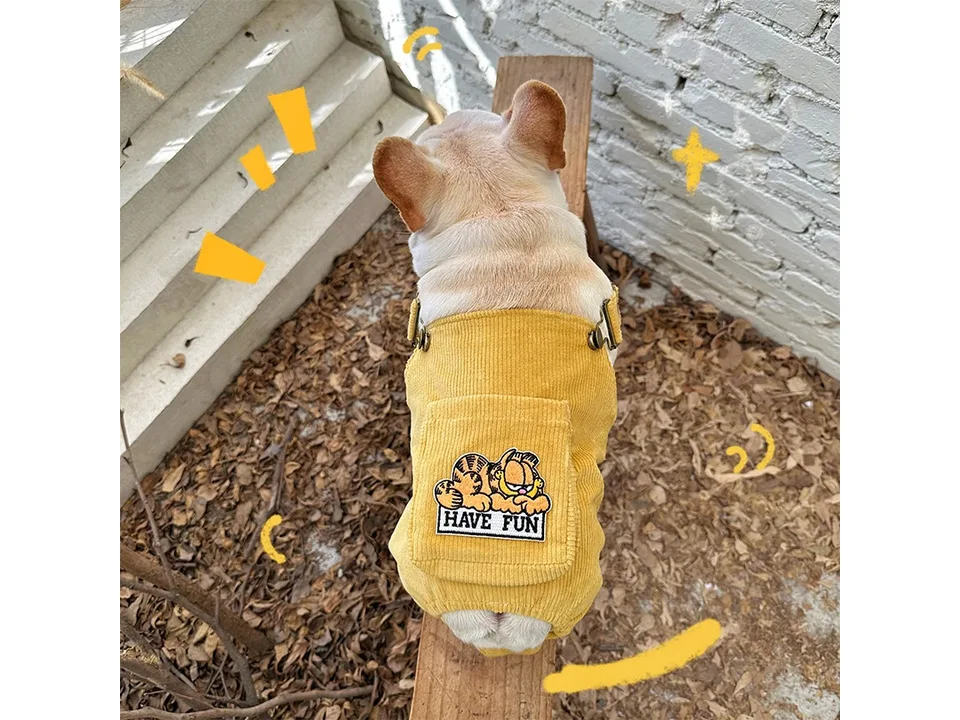 A French bulldog wearing a yellow corduroy jumpsuit with a Garfield patch on the back.
