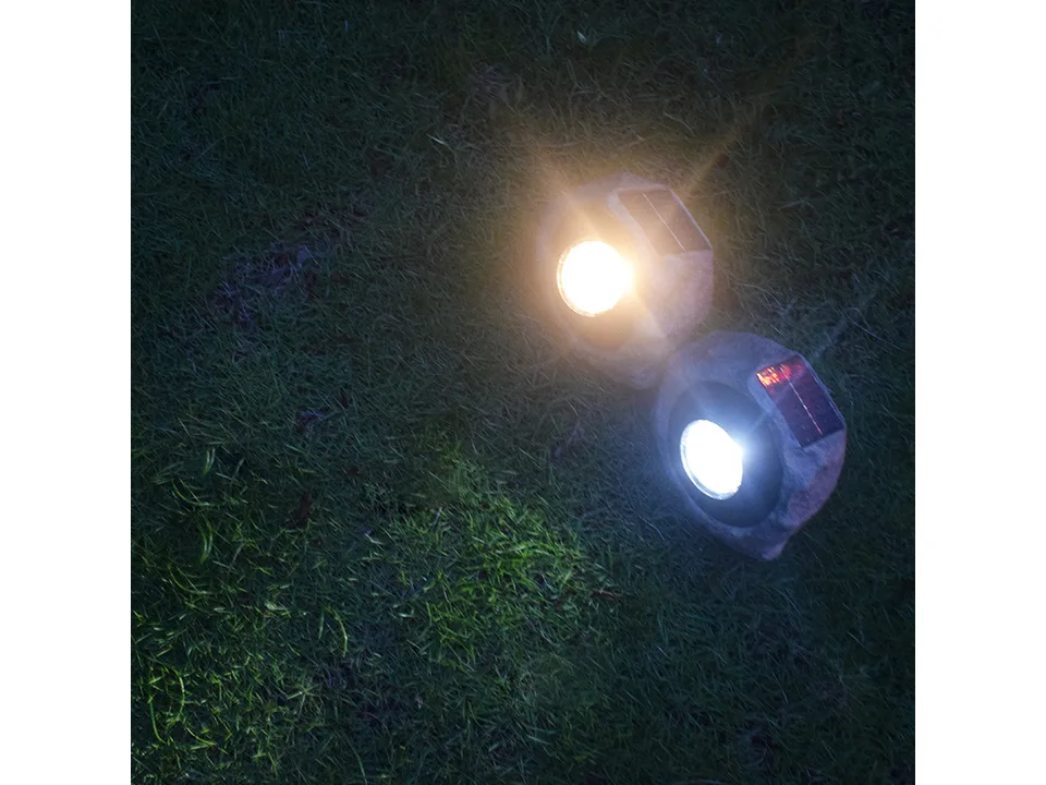 Two rock-shaped solar garden spotlights glowing on grass at night.