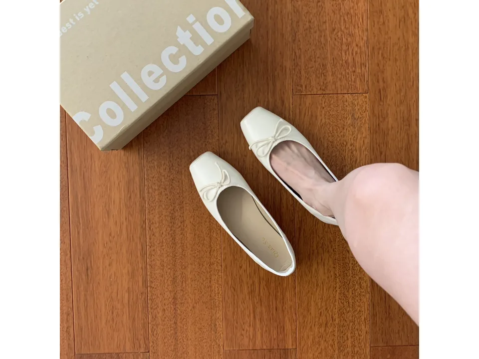 A pair of cream-colored square toe ballet flats with a bow, one being worn, next to a shoe box.