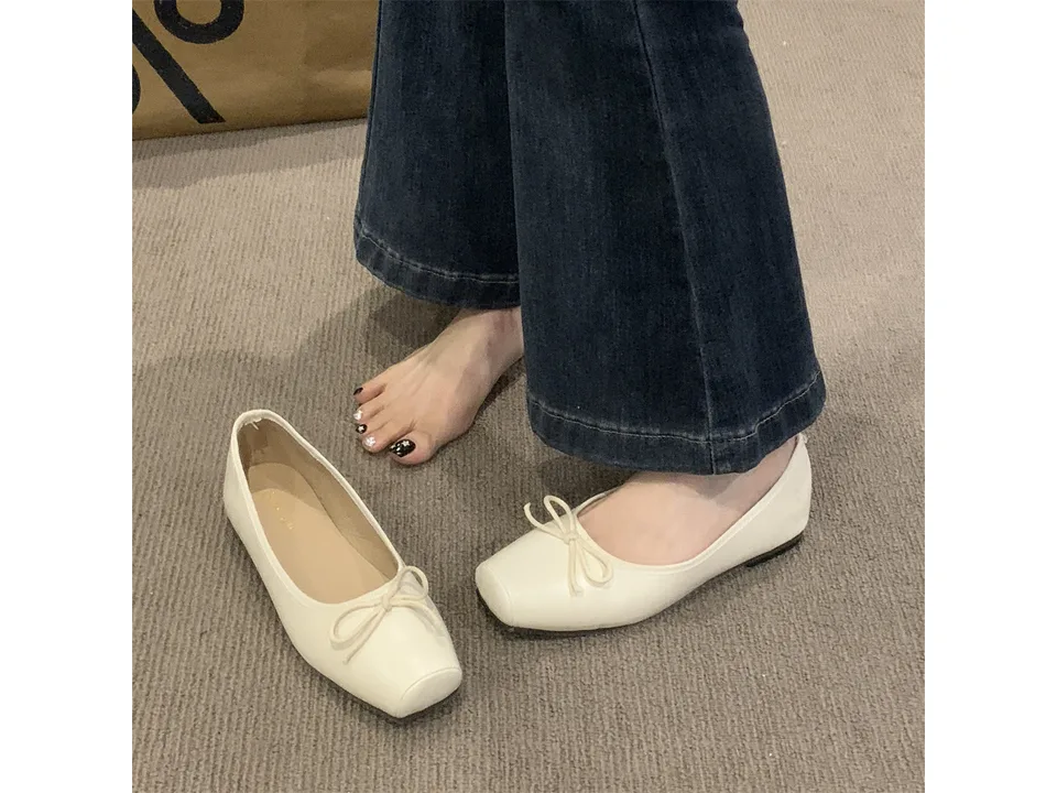 A pair of cream-colored square toe ballet flats with a bow, one being worn with jeans.