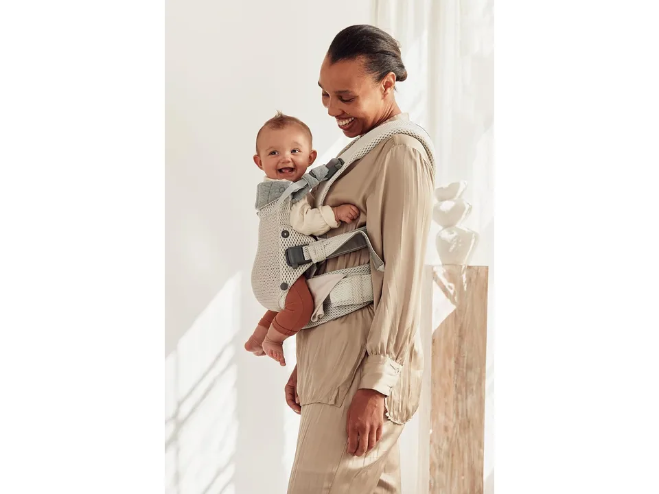 A woman wearing a beige baby carrier with a baby facing outward on her chest.