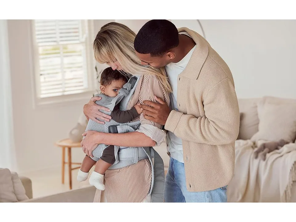 A woman wearing a grey baby carrier with a baby facing inward on her chest.