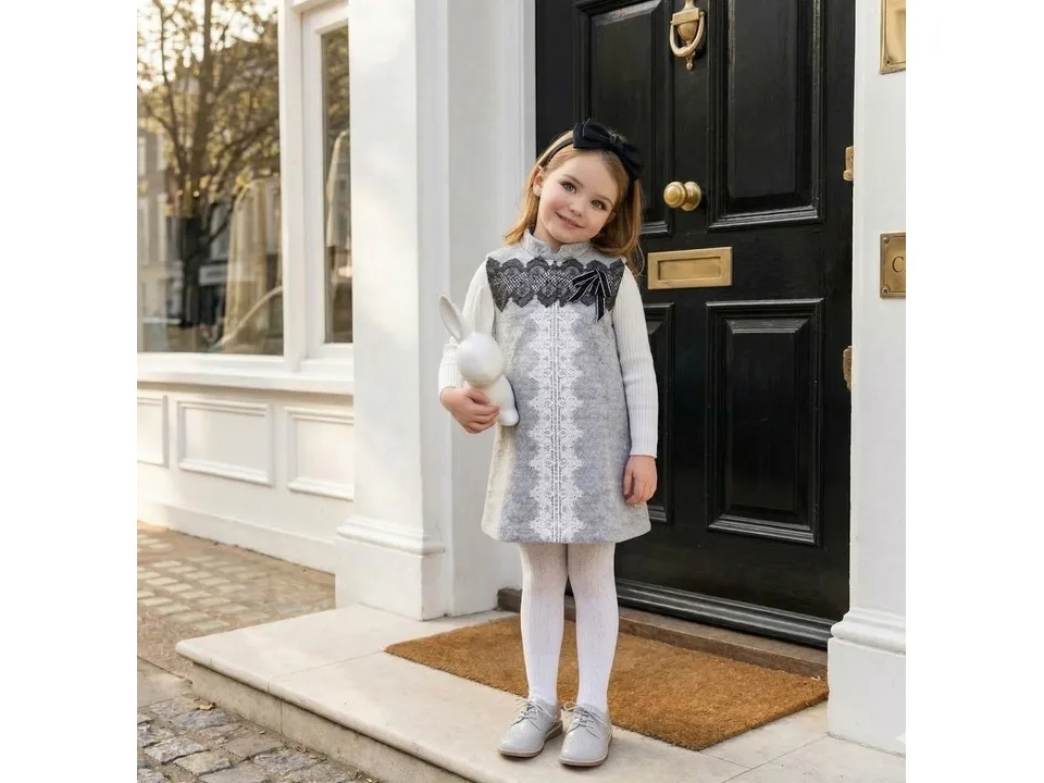 A young girl wearing the grey pinafore dress with a white long-sleeve top and white tights.