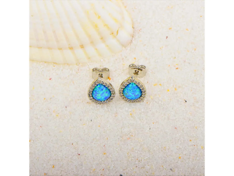 A pair of blue opal teardrop stud earrings resting on white sand next to a seashell.