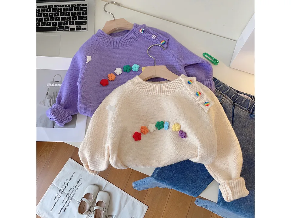 Two children's sweaters in purple and cream, featuring floral designs and shoulder buttons.