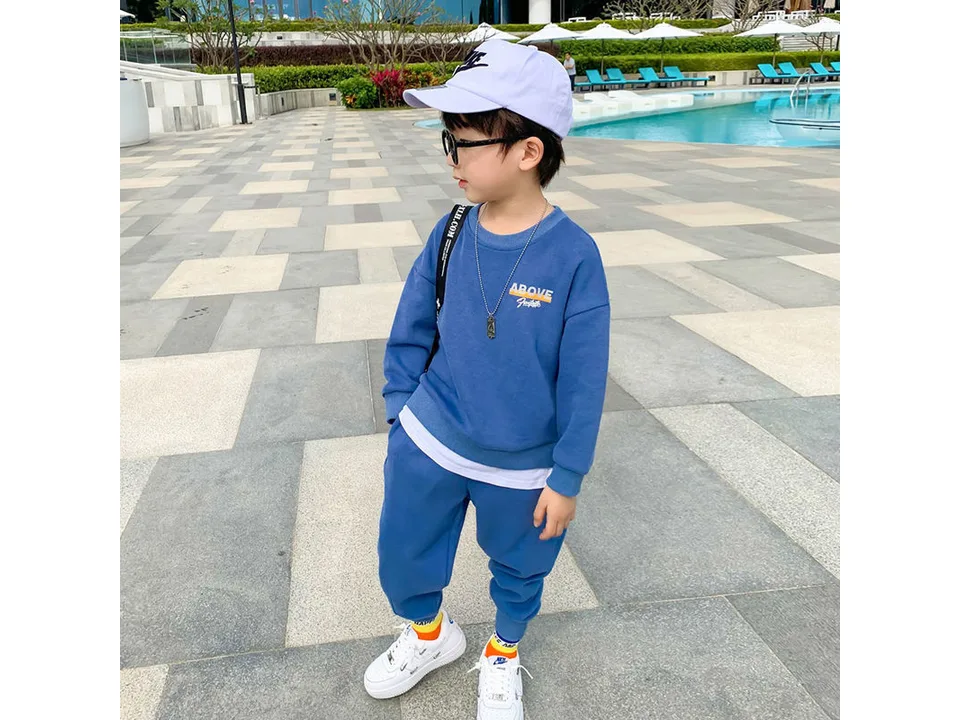 A young boy standing by a pool wearing a blue long-sleeve sweatshirt and matching jogger pants.
