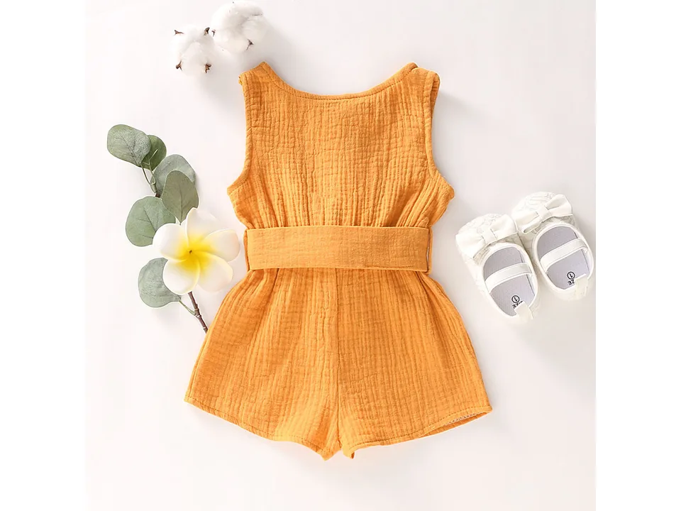Mustard yellow sleeveless toddler jumpsuit with a bow belt, displayed with decorative shoes and flowers.