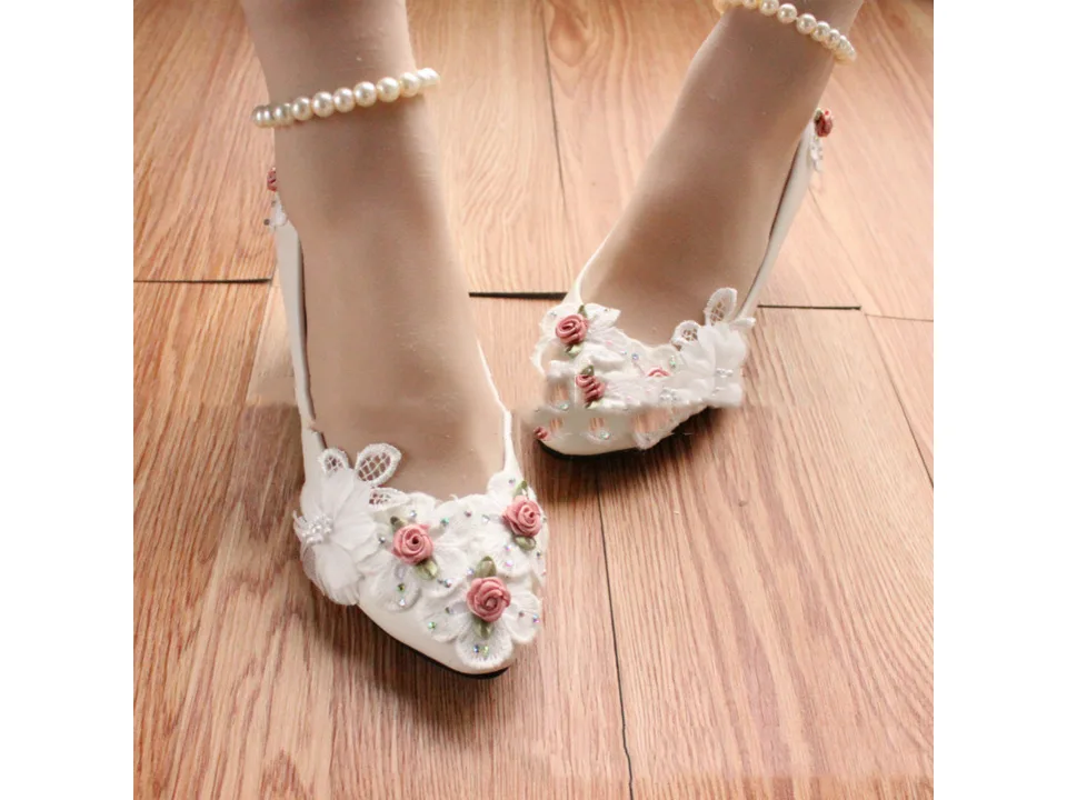 Pair of white wedding pumps with floral details and pearl anklets worn on feet.