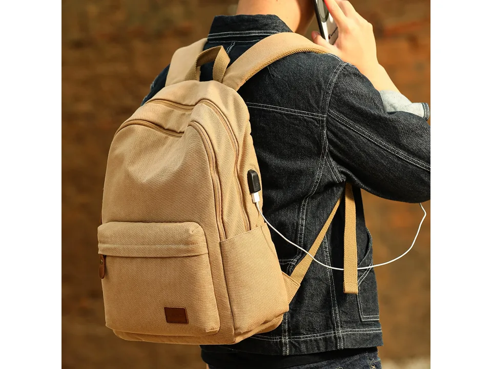 Person wearing the tan canvas backpack with a charging cable connected to the side port.